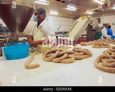 Die Arbeiter in einer Lebensmittelfabrik produzieren frische Würste, nutzen Industrieanlagen, um Därme zu befüllen, und handhaben Fleischprodukte manuell auf einem Stück Stockfoto