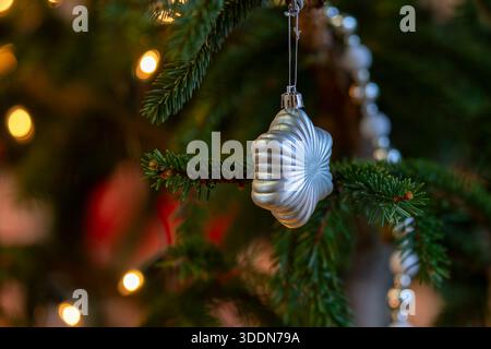 Elegantes silbernes Weihnachtsschmuck hängt an einem Tannenzweig mit warmen festlichen Lichtern im Hintergrund. Nahaufnahme einer silbernen Weihnachtsdekoration. Stockfoto
