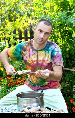 Ein Mann in einem Krawattenhemd sitzt in einem üppigen Garten und bereitet rohes Fleisch auf einem Spieß für ein Grillpicknick zu. Er hält einen Metallspieß über einem Topf, bereit für Gril Stockfoto