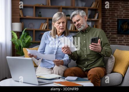 Seniorenpaar auf einem Sofa, gestresst und verwirrt, als sie Papierrechnungen mit Online-Banking auf einem Smartphone und Laptop vergleichen und mit unerwarteten Ausgaben und Budgetierung kämpfen Stockfoto