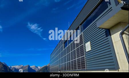 Blick auf das Gebäude mit in die Fassade integrierten Solarpaneelen vor dem Hintergrund der alpinen Berge. Nutzung erneuerbarer Energiequellen. Stockfoto