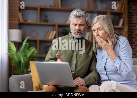 Seniorenpaar, das zu Hause auf einem Sofa sitzt und auf einen Laptop-Bildschirm blickt, mit Schock und Sorge, nachdem sie unerwartete schlechte Nachrichten, einen Betrug oder eine hohe Rechnung im Internet entdeckt haben Stockfoto