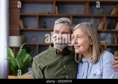 Glückliches Seniorenpaar, das mit einem Lächeln wegblickt, während es sich in seinem gemütlichen Wohnzimmer entspannt, symbolisiert eine liebevolle Beziehung und einen komfortablen Lebensstil im höheren Alter Stockfoto