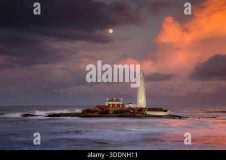 Ein einsamer Leuchtturm steht auf einer felsigen Insel, umgeben von einem Ozean mit stürzenden Wellen. Der Himmel zeigt atemberaubende Orange- und Violetttöne. Stockfoto