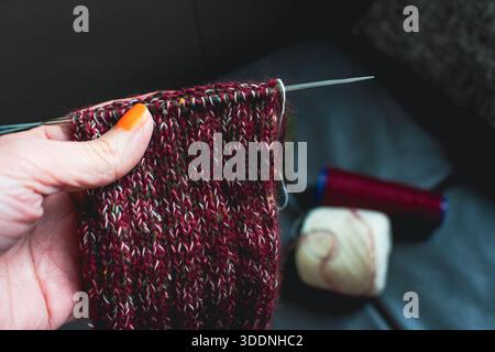 Frau, die kuschelige Stricksocken auf Nadeln in der Hand hält. Mischung aus rotem, grünem und weißem Garn auf Stricknadeln. Das Strickverfahren von Wollsocken. Stockfoto
