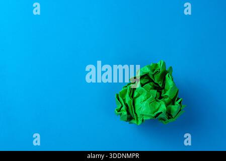 Grüne, zerknitterte Papierkugel auf blauem Hintergrund, auffällige abstrakte Textur für Recycling oder umweltfreundliche Ideen. Stockfoto
