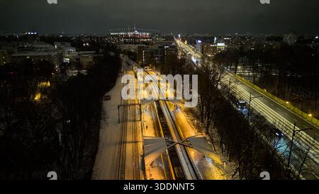 Verschneite Eisenbahnkorridor mit beleuchteten Bahnsteigen, Lichtpfaden von vorbeifahrenden Zügen und streifenden Autolichtern Warschau, Polen Stockfoto