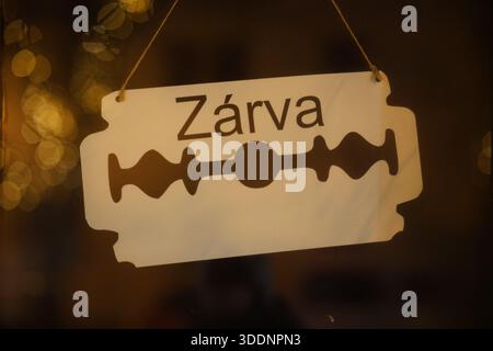Hängendes Schild „Zárva“ (geschlossen) in Form einer Rasierklinge in einem Schaufenster nachts in Eger, Ungarn. Eger, Ungarn Stockfoto