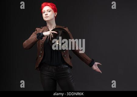 Stilvolle Frau mit kurzen roten Haaren in Steampunk Bowler Hut mit Schutzbrille auf Grau, macht einen Knick Stockfoto