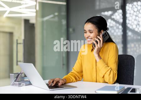Lächelnde Geschäftsfrau, die am Schreibtisch sitzt, aktiv auf dem Smartphone spricht, während sie auf dem Laptop schreibt, professionelle Aufgaben und Kommunikation in einer modernen Büroumgebung verwaltet Stockfoto