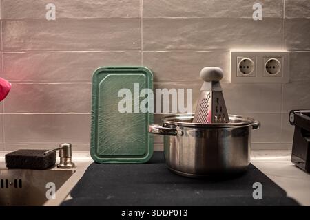 Küchenarbeitsplatte mit Edelstahltopf, Metallreibe innen, grünes Schneidebrett aus Kunststoff und Waschbecken. Tägliche Kochszene in einem modernen Zuhause Stockfoto