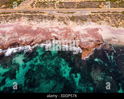 Ein Blick aus der Vogelperspektive zeigt die zerklüftete rote Felsküste und die türkisfarbenen Wellen des Ozeans. Die ruhige Landschaft bietet stürzende Wellen und eine gewundene Straße. Stockfoto