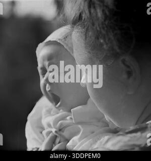 Slawjansk, ukrainische SSR, Sowjetunion, Ende der 1960er Jahre Ein ehrliches Archivbild einer Mutter, Kravelis Valentina Timofejewna, die ihren gähnenden Sohn Kravelis Dmitri Michailowitsch in der Hand hält. Das Baby trägt eine traditionelle Baumwollhaube mit Spitzenbesatz. Die Mutter sieht ihr verschlafenes Kind vor einem weichen, verschwommenen Hintergrund zärtlich an. Dieses authentische Schwarzweiß-Vintage-Foto fängt einen lustigen natürlichen Moment der Müdigkeit und der Atmosphäre des Familienlebens in der friedlichen industriellen Donbass-Region während der UdSSR-Ära ein. Stockfoto