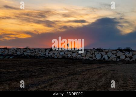 Silhouette einer Person, die während eines farbenfrohen Sonnenuntergangs mit Wolken auf einem felsigen Wellenbrecher steht. Stockfoto