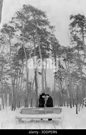 Slawjansk, ukrainische SSR, Sowjetunion, Anfang der 1950er Jahre Eine romantische vertikale Winterszene im Park des berühmten Slaviansk Resorts. Ein liebevolles Paar, genannt Kravelis Michail Iljitsch und Kravelis Valentina Timofeevna, steht hinter einer Holzbank, die mit tiefem Schnee bedeckt ist. Sie reden leise zwischen hohen, majestätischen Kiefern. Im Hintergrund sind weiße Spalten sichtbar. Dieses authentische Vintage-Schwarz-weiß-Foto fängt einen Moment der Intimität und der friedlichen Atmosphäre der industriellen Donbass-Region während der Nachkriegszeit der UdSSR ein. Stockfoto
