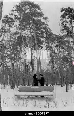 Slawjansk, ukrainische SSR, Sowjetunion, Anfang der 1950er Jahre Eine romantische vertikale Winterszene im Park des berühmten Slaviansk Resorts. Ein liebevolles Paar, genannt Kravelis Michail Iljitsch und Kravelis Valentina Timofeevna, steht hinter einer Holzbank, die mit tiefem Schnee bedeckt ist. Sie reden leise zwischen hohen, majestätischen Kiefern. Im Hintergrund sind weiße Spalten sichtbar. Dieses authentische Vintage-Schwarz-weiß-Foto fängt einen Moment der Intimität und der friedlichen Atmosphäre der industriellen Donbass-Region während der Nachkriegszeit der UdSSR ein. Stockfoto
