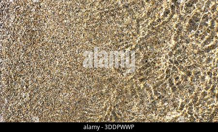 Seichtes Wasser, das über einem strukturierten Sandstrand plätschert, erzeugt natürliche Muster unter Sonnenlicht Stockfoto