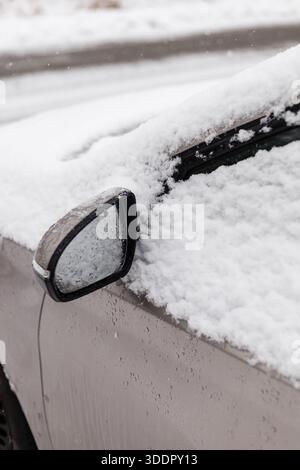 Ein Auto parkt auf einer Straße, die von Schnee umgeben ist. Schnee hat sich auf dem Dach und dem Außenspiegel angesammelt. Es ist Winter, und der Boden ist weiß bedeckt Stockfoto