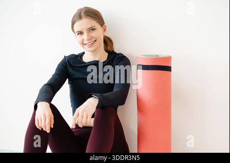 Eine Frau mit einem Pferdeschwanz und einem schwarzen Langarmhemd sitzt auf dem Boden und eine rosafarbene Yogamatte neben ihr. Sie sieht entspannt und glücklich nach einem Workou aus Stockfoto
