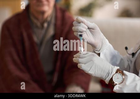 Kaukasischer Seniorenmann sitzt im Hintergrund, während der Arzt Handschuhe trägt, die Spritze mit Medikamenten vorbereitet, Schwerpunkt auf der Gesundheitsversorgung und der Pflege älterer Patienten Stockfoto