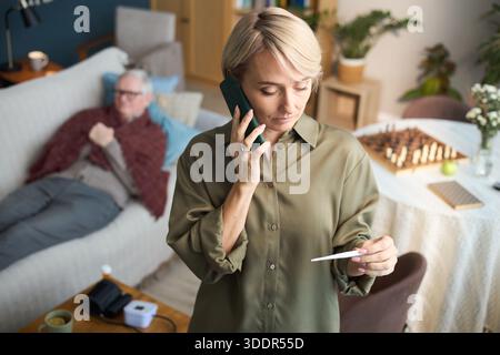 Kaukasische Frau mittleren Alters, die mit dem Smartphone spricht, während sie ein Thermometer hält, ein älterer kaukasischer Mann, der im Hintergrund auf der Couch liegt, medizinisches Gerät und Schachbrett sichtbar auf dem Tisch Stockfoto