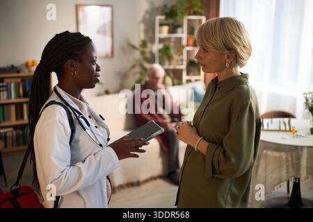 Schwarze junge Erwachsene Frau Doktor hält ein digitales Tablet, spricht mit einer kaukasischen Frau mittleren Alters im Wohnzimmer, während ein älterer kaukasischer Mann auf einem Sofa im Hintergrund sitzt Stockfoto