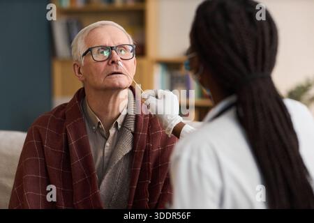 Kaukasischer Senior Mann sitzend, während schwarze Frau Gesundheitspersonal, die Nasentupfer-Test, beide drinnen, Mann mit Brille und Blick nach oben, medizinische Untersuchung im Gange Stockfoto
