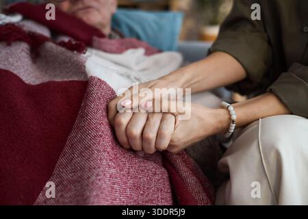 Kaukasische Frau mittleren Alters, die Hand eines älteren kaukasischen Mannes hält, der unter der Decke liegt, Unterstützung und Komfort zeigt, Nahaufnahme der Hände, die Sorgfalt und Mitgefühl betonen Stockfoto
