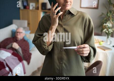Kaukasische Frau mittleren Alters, die ein Thermometer hält, während sie mit dem Smartphone spricht, im Wohnzimmer mit einem älteren kaukasischen Mann, der auf der Couch unter einer Decke im Hintergrund liegt Stockfoto