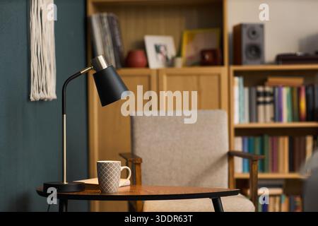 Moderne Tischlampe, die auf einem hölzernen Beistelltisch neben einem gemusterten Kaffeetasse in einer gemütlichen Leseecke steht, mit Büchern und gerahmten Fotos im Hintergrund Stockfoto