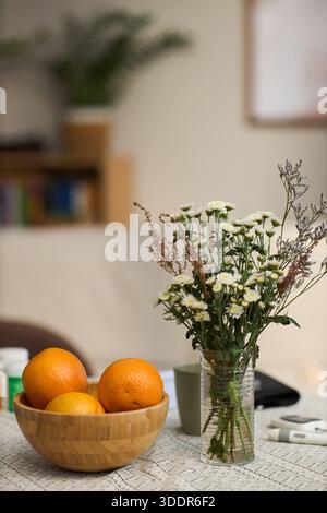 Stillleben mit Glasvase mit Gänseblümchen und Wildblumen, die neben einer mit Orangen gefüllten Holzschale auf einem Tischtuch aus Spitze stehen, verschwommener Hintergrund deutet auf eine Inneneinrichtung hin Stockfoto