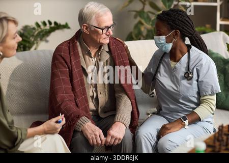 Senior-kaukasischer Mann sitzt auf einem Sofa mit Decke bedeckt und erhält Unterstützung von der schwarzen Krankenschwester mit medizinischen Peelings und Maske, während kaukasische Frau mittleren Alters beobachtet Stockfoto