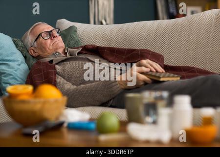 Älterer kaukasischer Mann, der mit geschlossenen Augen auf dem Sofa liegt, eine Brille trägt und ein Buch in der Hand hält, Arzneiflaschen und Obst im Vordergrund auf dem Tisch, was auf eine Heimerneuerung hindeutet Stockfoto