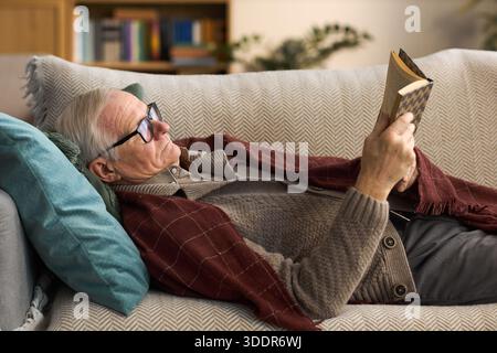Älterer kaukasischer Mann, der auf dem Sofa liegt und Buch liest, eine Brille trägt und sich mit Kopf auf Kissen, Bücherregal und Pflanzen im Hintergrund entspannt und die Freizeitaktivität zu Hause zeigt Stockfoto