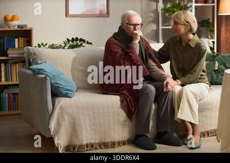 Ein älterer kaukasischer Mann sitzt auf dem Sofa, der den Mund mit der Hand bedeckt, während die junge Erwachsene kaukasische Frau neben ihm sitzt und tröstet und unterstützt während eines schwierigen Moments Stockfoto