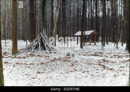 Ein handgefertigter, schlanker Unterschlupf, der aus Holzästen gegen einen Baumstamm in einem schneebedeckten Laubwald gebaut wurde. Stockfoto