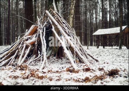 Ein handgefertigter, schlanker Unterschlupf, der aus Holzästen gegen einen Baumstamm in einem schneebedeckten Laubwald gebaut wurde. Stockfoto