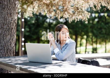 Eine konzentrierte junge Frau in einem blau gestreiften Hemd sitzt mit einem Laptop an einem Holztisch und genießt das natürliche Licht und die Außenatmosphäre eines Cafés Stockfoto