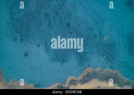 Faszinierende Draufsicht einer gefrorenen blauen Seenoberfläche, die abstrakten Kunstwerken ähnelt. Komplizierte Eismuster, eingeschlossene Blasen und Froststrukturen stehen im Kontrast zur Sandküste und schaffen einen atemberaubenden kalten Winterhintergrund mit Kopierraum. Stockfoto