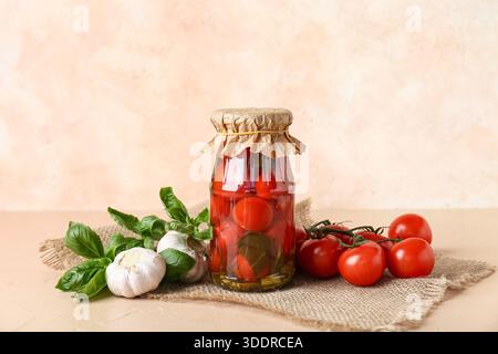 Glas mit leckeren Dosentomaten mit Knoblauch und Basilikumblättern auf beigem Hintergrund Stockfoto