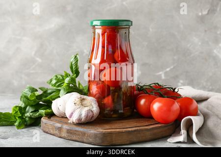 Glas mit leckeren Dosentomaten mit Knoblauch und Basilikumblättern auf grauem Hintergrund Stockfoto