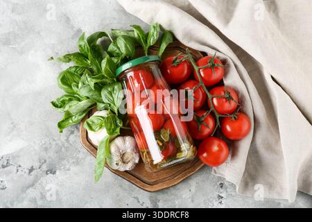 Glas mit leckeren Dosentomaten mit Knoblauch und Basilikumblättern auf grauem Hintergrund Stockfoto