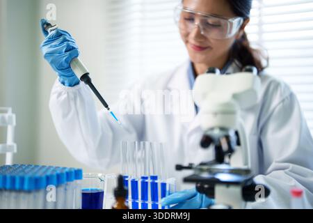 Erwachsene asiatische Forscherin pipettiert präzise blaue Lösung in Laborröhrchen Sie trägt eine weiße Schutzbrille und blaue Handschuhe wo Stockfoto