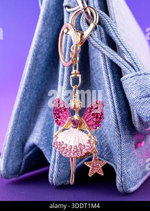 Nahaufnahme eines hellen Schlüsselanhängers aus Metall in Form einer Fee mit Flügeln aus rosa Strasssteinchen auf einer blauen Tasche Stockfoto