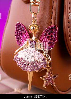 Nahaufnahme eines hellen Schlüsselanhängers aus Metall in Form einer Fee mit Flügeln aus rosa Strasssteinchen auf einer braunen Tasche Stockfoto