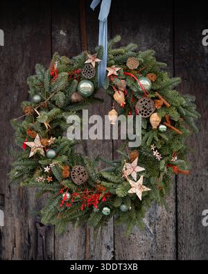 Handgefertigter Weihnachtskranz aus frischen immergrünen Zweigen, verziert mit natürlichen Ornamenten und hängt an einer rustikalen Holztür. Stockfoto