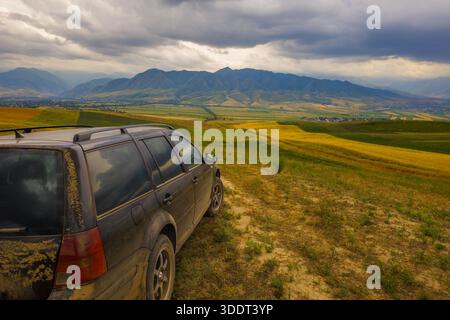 Ein staubbedecktes Auto parkt auf einer schroffen Feldstraße und bietet einen atemberaubenden Blick auf ein ruhiges Tal. Der bewölkte Himmel und die bergige Landschaft vermitteln ein Gefühl von Abenteuer und Ruhe. Stockfoto