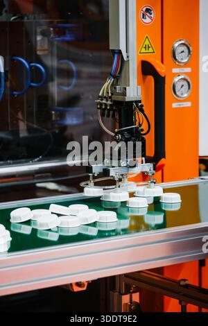Automatische Verschließmaschine Robotergreifer, die weiße Kunststoffflaschenverschlüsse vom Förderband in Hochgeschwindigkeitsverpackungslinien aufnimmt Stockfoto