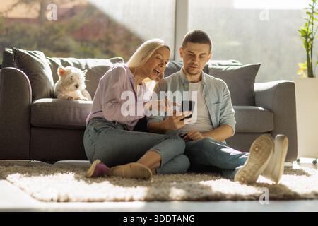 Junge Paare sitzen zu Hause auf dem Boden und teilen Inhalte auf einem Smartphone Stockfoto