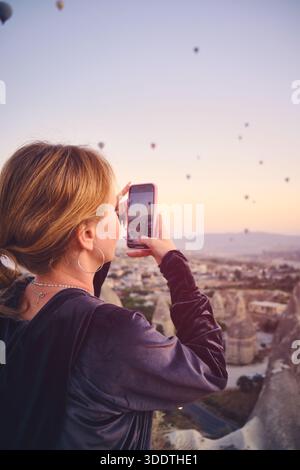 Die junge Frau fängt den malerischen Blick auf die bunten Heißluftballons ein, die bei Sonnenaufgang über die dramatische felsige Landschaft Kappadokiens aufsteigen Stockfoto
